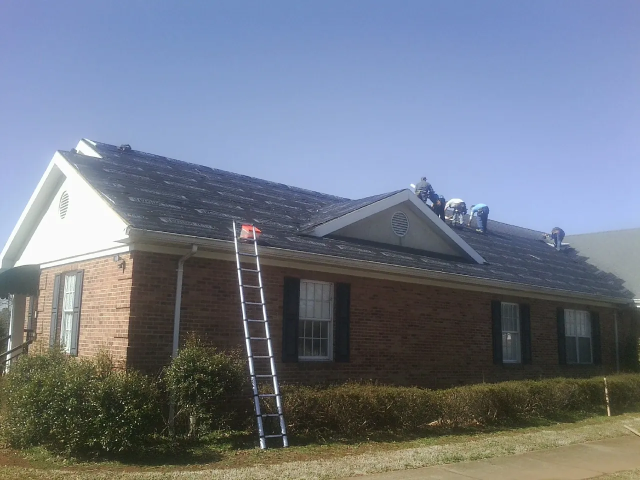 Crew tearing shingles off a brick ranch roof with a ladder set — Metal Roof Repair work by our crew in Gladbrook