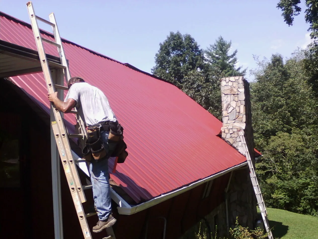 Roofer on a ladder beside a red metal roof and stone chimney — Soffit & Fascia Repair by Roofing Brothers in Gladbrook